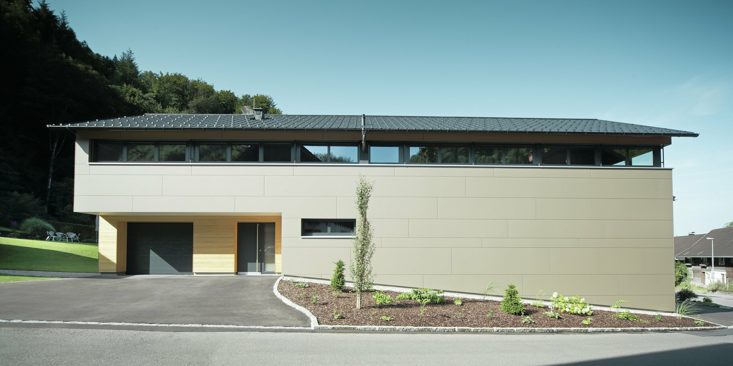 Das Einfamilienhaus in Hohenems präsentiert sich in einer modernen und stilvollen Fassade aus PREFA Aluminium Verbundplatten in Bronzemetallic, mit einem darüberliegenden Dach aus PREFA Dachplatten in P.10 Anthrazit. Die klare Linienführung und das durchdachte Farbkonzept des Designs integrieren das Gebäude nahtlos in die natürliche Umgebung. Die Gestaltung zeigt die Kombination aus Funktionalität und Ästhetik, die PREFA Produkte für zeitgemäße Wohnarchitektur bieten.