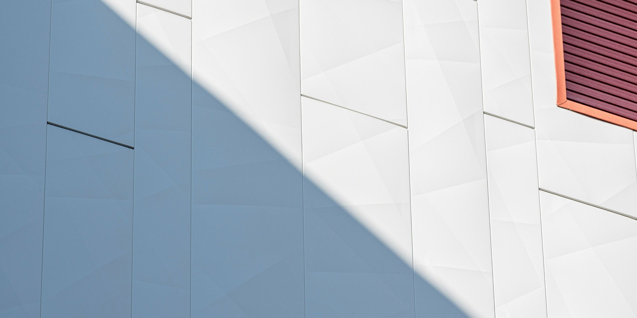 Detailaufnahme der sanierten Fassade der Wohnanlage Alba B mit PREFA Siding.X Aluminiumfassadenpaneelen: Großformatige, vertikal verlegte Paneele mit feinen Kantungen erzeugen eine lebendige, geometrische Struktur. Ein schräg einfallender Schatten betont die metallische Oberfläche und sorgt für eindrucksvolle Licht- und Schattenspiele. Am rechten Bildrand setzt eine rot akzentuierte Fensterlaibung einen starken Kontrast. Die PREFA Aluminiumfassade überzeugt durch hohe Widerstandsfähigkeit, Langlebigkeit und eine dauerhaft wartungsarme, ästhetische Gebäudehülle.