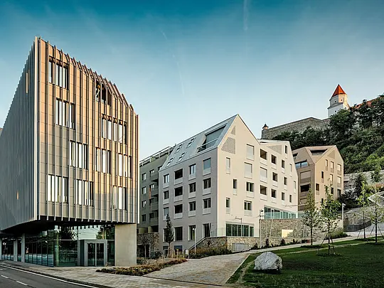 Markantes Zusammenspiel aus moderner Architektur und historischem Kontext im Stadtquartier Vydrica in Bratislava: Im Vordergrund fasziniert ein skulpturaler Baukörper mit vertikaler Fassadenstruktur, während dahinter steil geneigte Dächer mit PREFA Aluminium Dachschindeln in Prefaweiß und Sandbraun präzise Akzente setzen. Die klar gegliederten Dachflächen mit integrierten Fenstern bilden einen starken visuellen Kontrast zur Burgkulisse am Hang. PREFA Aluminium überzeugt durch hohe Formbarkeit, Langlebigkeit und extreme Witterungsbeständigkeit – ideal für architektonisch anspruchsvolle Stadtprojekte.