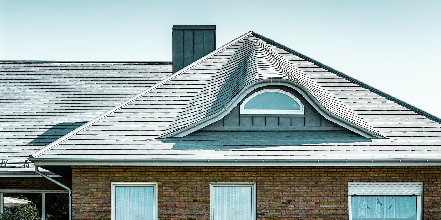 Einfamilienhaus aus den 70ern mit einem Schindeldach aus Aluminium in P.10 Steingrau mit geschwungener Gaube mit rundem Fenster