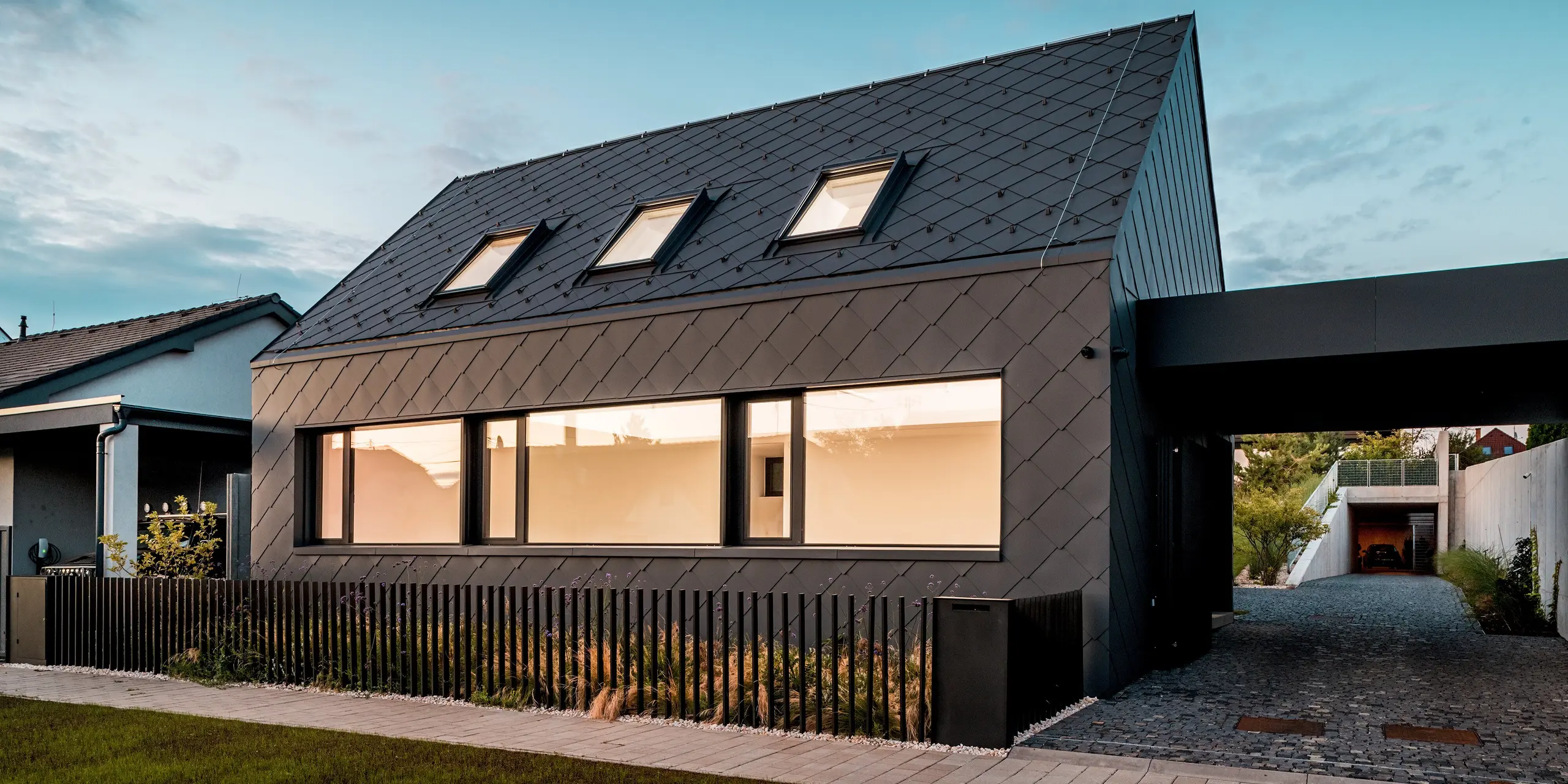 L'image présente une maison individuelle moderne à Trnava-Modranka au crépuscule, entièrement revêtue de losanges de toiture et de façade 44 x 44 PREFA, coloris P.10 gris sombre. La lumière des fenêtres crée un contraste chaleureux avec le revêtement en aluminium sombre à la texture uniforme. La disposition précise des losanges souligne la pureté des lignes du bâtiment et met en valeur la qualité et la robustesse des matériaux. Durables, les produits en aluminium PREFA offrent une protection optimale contre les intempéries et confèrent au bâtiment une allure remarquable et intemporelle.