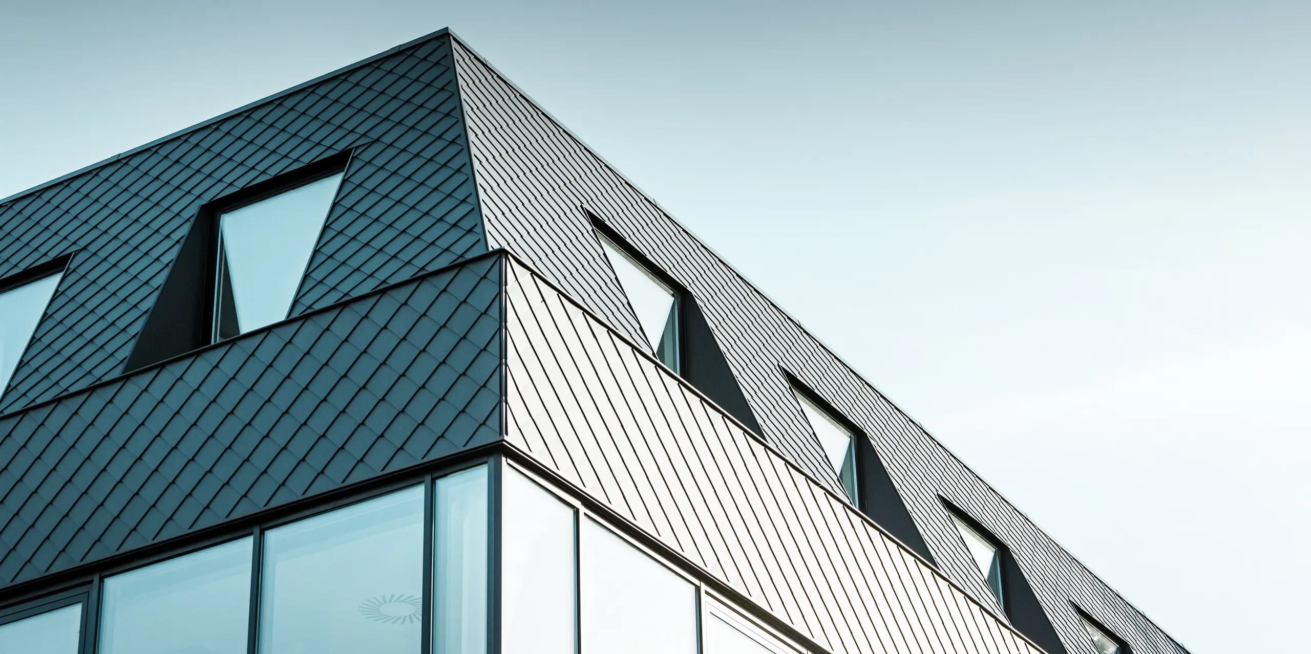 Das moderne Bürogebäude Kasteeltje Heizel in Strombeek-Bever nahe Brüssel verfügt über eine markante PREFA Aluminium-Fassade aus anthrazitfarbenen Wandrauten. Die Nahaufnahme der geometrischen Rautenstruktur bildet einen spannenden Kontrast zu den großen Glasflächen und verleiht dem Gebäude eine dynamische, futuristische Ausstrahlung. Die langlebigen und wartungsarmen PREFA Aluminiumprodukte vereinen Ästhetik, Nachhaltigkeit und Funktionalität in einem architektonischen Eyecatcher.