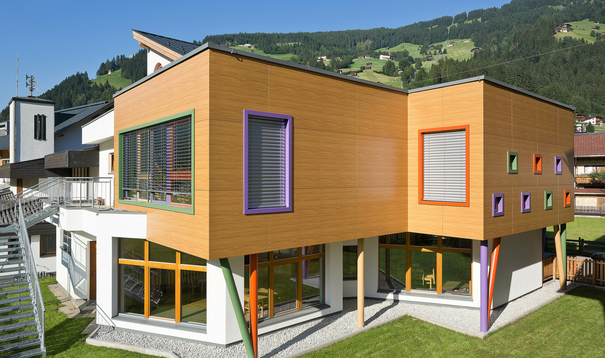 Modernes, zweigeschossiges Gebäude mit auffälliger Holzoptik-Fassade und farbenfrohen Akzenten. Fensterrahmen in Grün, Lila und Orange setzen lebendige Kontraste, während schräge, bunte Stützen die obere Etage tragen. Das Gebäude steht auf einer gepflegten Rasenfläche in einer alpinen Umgebung mit grünen Hügeln und Häusern im Hintergrund.