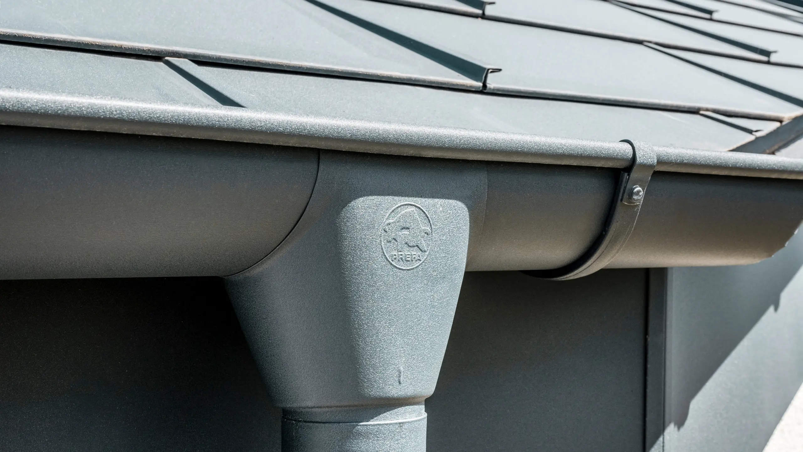 Nahaufnahme eines Hausdaches mit PREFA Dachplatte R.16 in Anthrazit sowie detailreicher Ansicht der farblich abgestimmten PREFA Dachrinne und Fallrohr aus Aluminium bei sonnigem Wetter.
