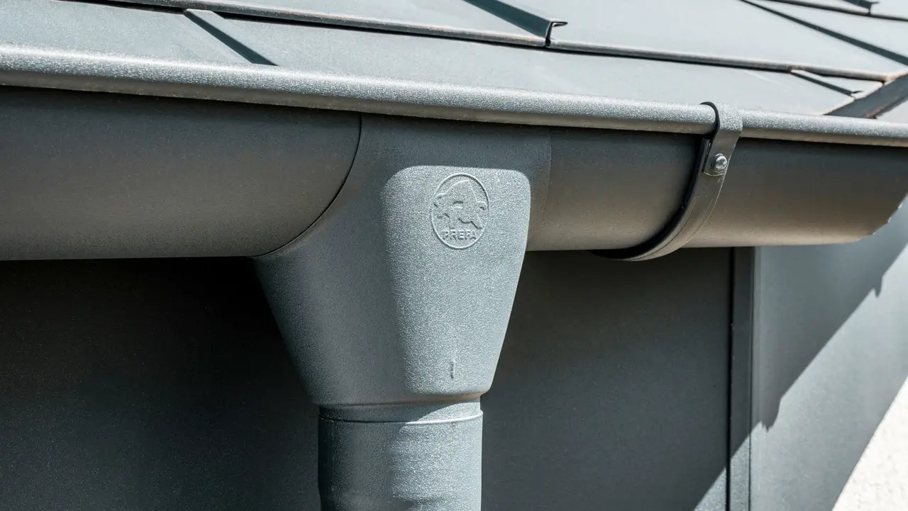 Nahaufnahme eines Hausdaches mit PREFA Dachplatte R.16 in Anthrazit sowie detailreicher Ansicht der farblich abgestimmten PREFA Dachrinne und Fallrohr aus Aluminium bei sonnigem Wetter.