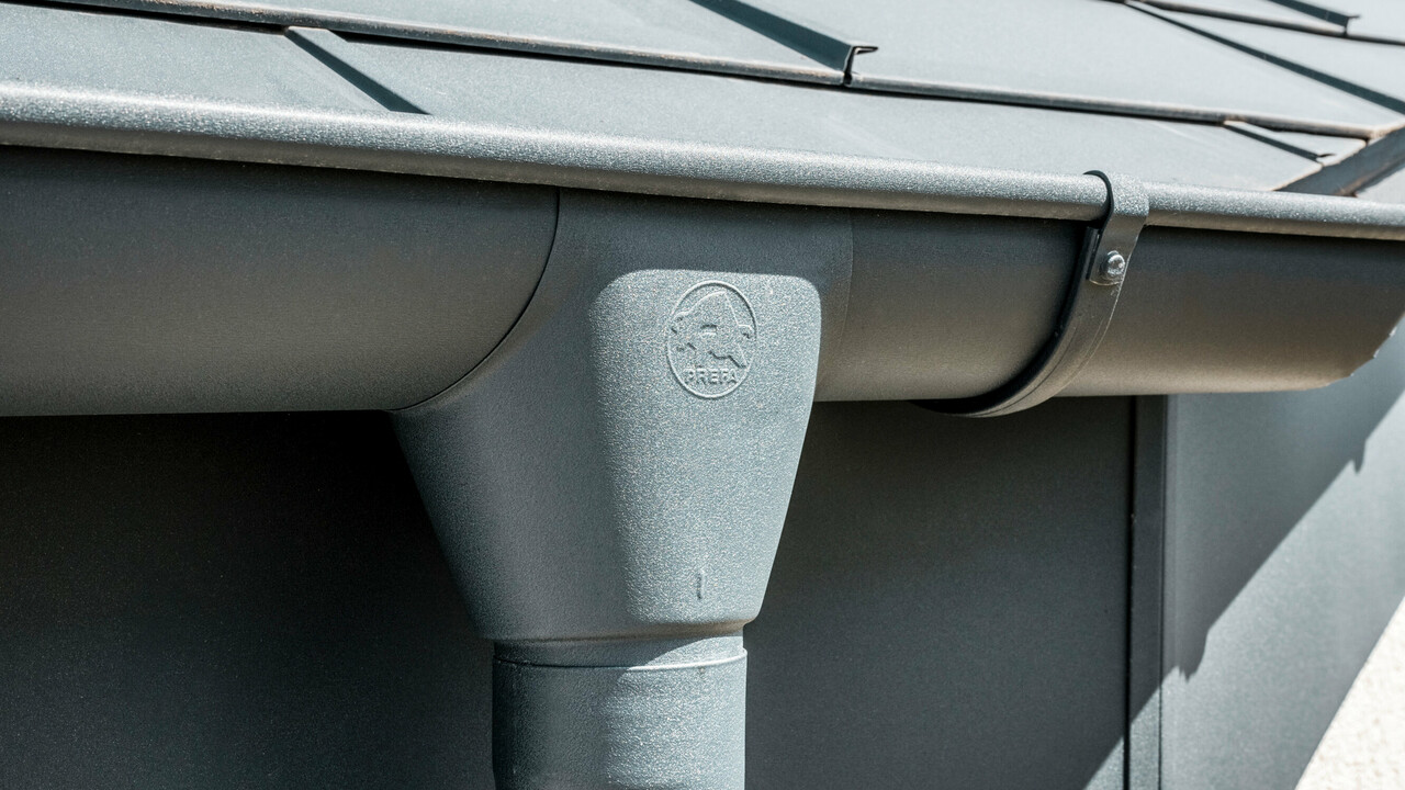 Nahaufnahme eines Hausdaches mit PREFA Dachplatte R.16 in Anthrazit sowie detailreicher Ansicht der farblich abgestimmten PREFA Dachrinne und Fallrohr aus Aluminium bei sonnigem Wetter.