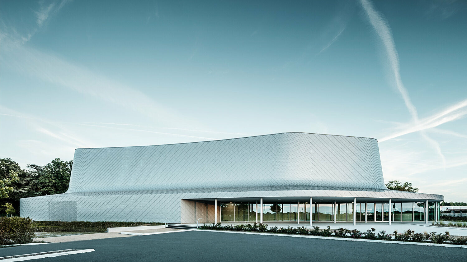 Vue latérale du bâtiment incluant le parvis. Le vitrage de la zone d'entrée donne l'impression que le bâtiment est partiellement transparent.