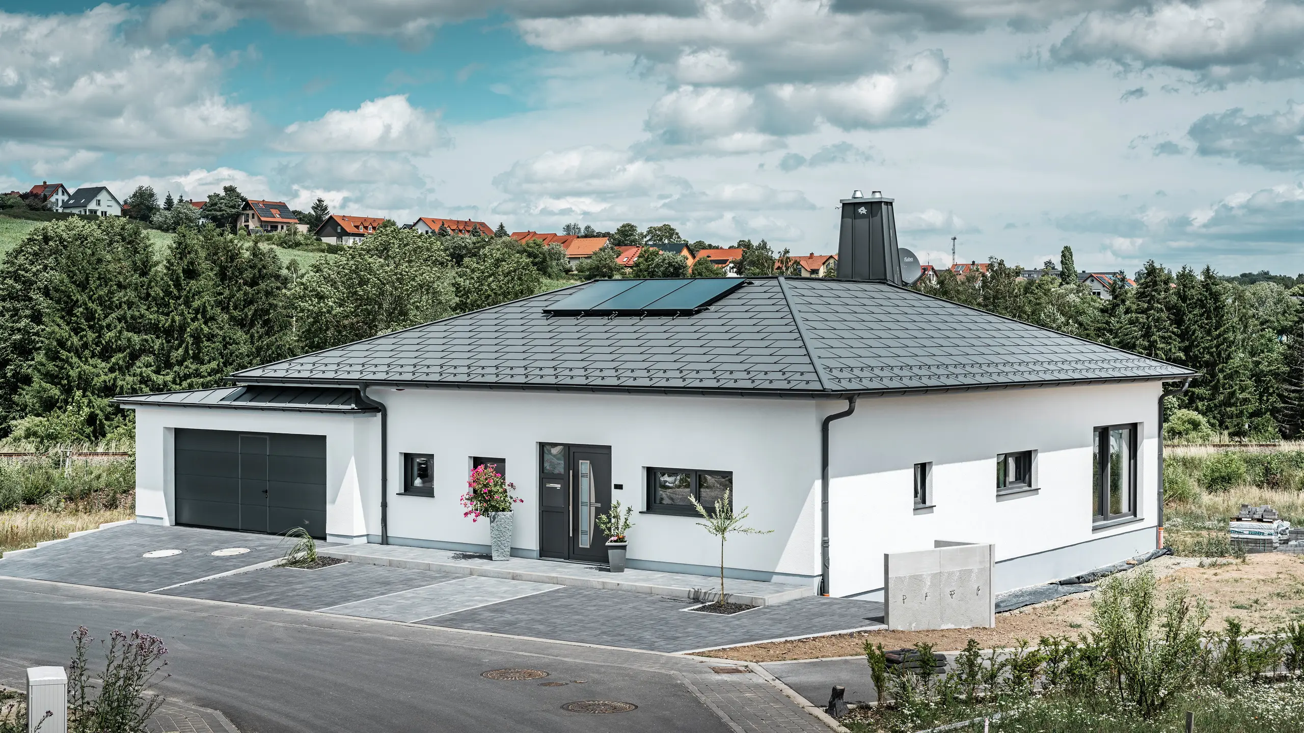 Elegante eengezinswoning in Ilmenau, Thüringen, met een hoogwaardig PREFA-dak in de kleur P.10 donkergrijs, voorzien van de robuuste dakpan R.16. De garage sluit qua kleur en stijl aan door het gebruik van PREFALZ. De blikvanger van het pand is het functioneel ontworpen atrium en de met de PREFA-stier versierde schoorsteen, die het moderne design en de innovatieve daktechnologie van PREFA onderstrepen. De zonnepanelen passen naadloos in het duurzame concept van het huis. Het doordachte totaalconcept van de bungalow wordt gecompleteerd door het PREFA-dakafvoersysteem, dat eveneens in donkergrijs is uitgevoerd.