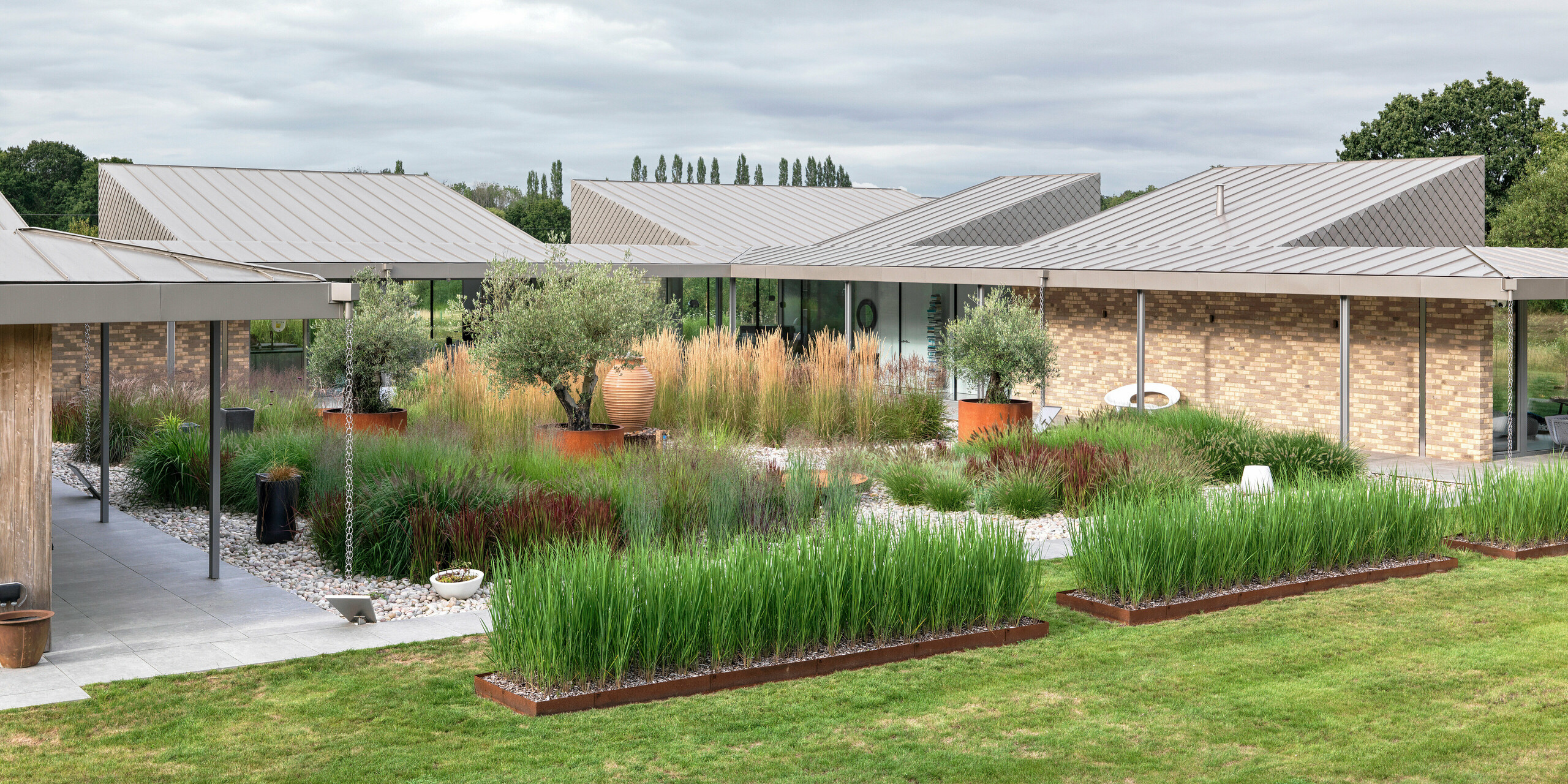 Vue panoramique sur le jardin d'un bungalow moderne du West Sussex, doté d'une toiture multi-sections en aluminium PREFALZ couleur P.10 bronze. Le point central est une cour intérieure naturelle, agrémentée de graminées, d'oliviers et d'éléments en terre cuite, entourée de passerelles couvertes aux supports délicats. Les lignes épurées du toit, en aluminium PREFA durable, soulignent la structure architecturale tout en offrant une résistance maximale aux intempéries et un entretien minimal.