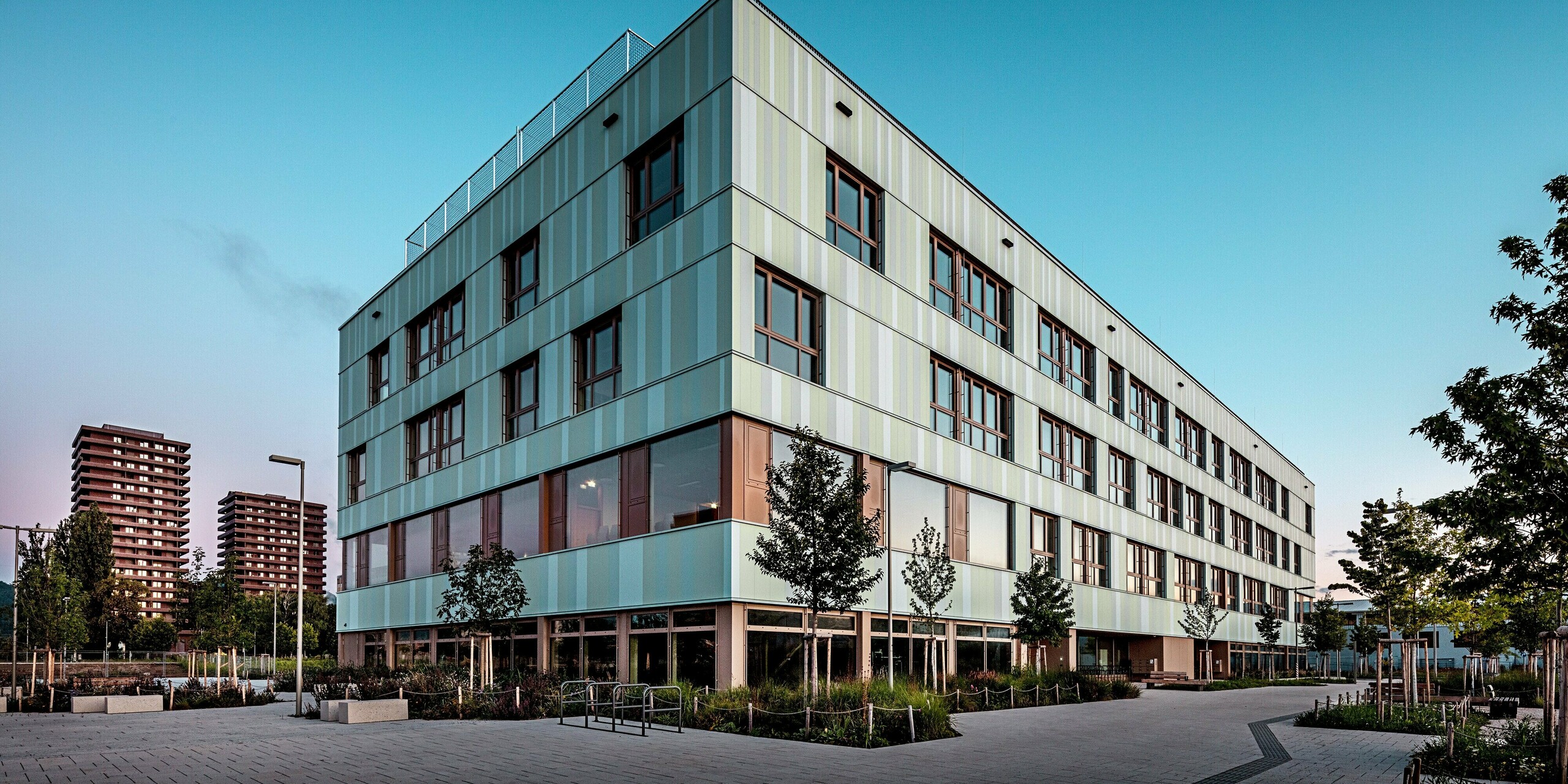 Die moderne und ansprechend gestaltete Fassade der AHS Reininghaus in Graz fällt durch die Verwendung von PREFA-Sidings in Sonderfarben auf. Die vertikal angeordneten Aluminiumpaneele in sanften Grüntönen und Weiß verleihen dem Gebäude eine elegante und zugleich dynamische Ausstrahlung. Die Fensterrahmen in warmem Braun bilden einen gelungenen Kontrast zur helleren Fassade und schaffen eine harmonische Verbindung. Im Vordergrund des Bildes sind gut gepflegte Grünflächen und Bäume zu sehen, die das moderne Design des Gebäudes perfekt ergänzen. Der Außenbereich ist mit Wegen und Fahrradbügeln ausgestattet, was die Funktionalität und Nachhaltigkeit des Projekts unterstreicht. Die Schule fügt sich harmonisch in ihre urbane Umgebung ein und setzt durch ihre Architektur und Materialwahl ein klares Statement.