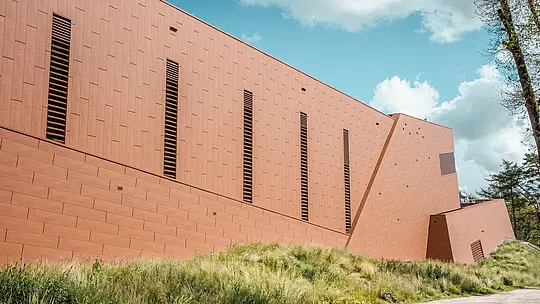 Vue latérale saisissante de la station de pompage d'eau potable WML à Lottum, mettant en valeur un bâtiment allongé à la structure bien définie, niché au cœur d'une prairie naturelle. La façade en aluminium PREFA Siding.X, à l'aspect chaleureux et rouillé, est structurée par de fines persiennes disposées de manière rythmée, créant un jeu d'ombre et de lumière captivant. La surface finement facettée offre un rendu dynamique et sophistiqué sous différents angles. Légers, durables et entièrement recyclables, les panneaux en aluminium PREFA garantissent une excellente résistance aux intempéries, une tenue des couleurs optimale et une enveloppe de bâtiment durable et nécessitant peu d'entretien.