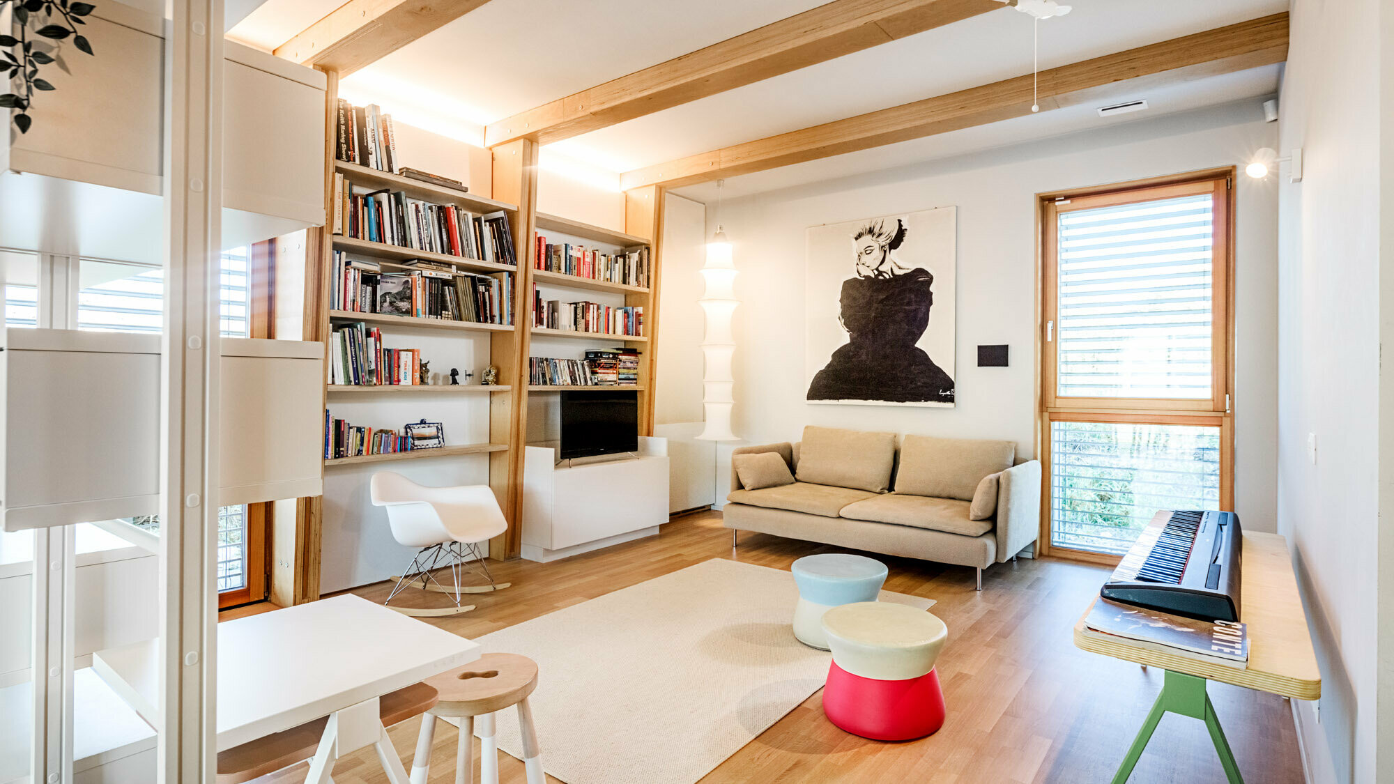 Photo de l’intérieur, les murs blancs sont rehaussés d’une bibliothèque et d’un tableau. La pièce contient aussi un canapé, un clavier électronique, des tabourets, une table et un fauteuil.