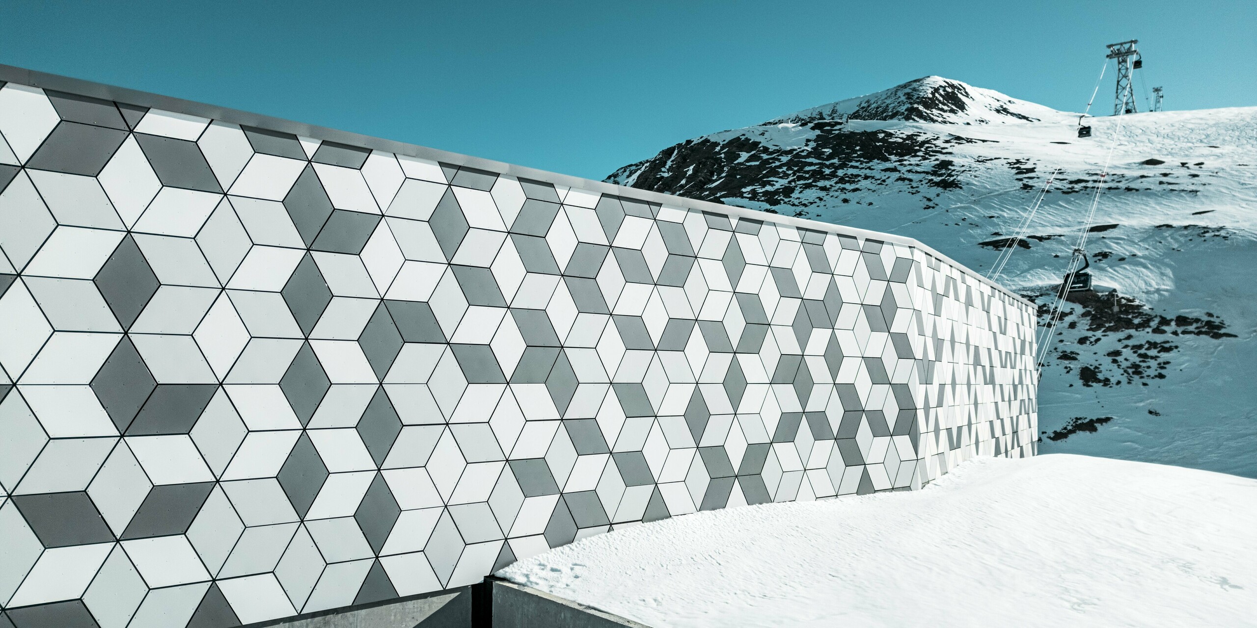La façade de la station de téléphérique des Deux Alpes est un chef-d'œuvre d'architecture moderne. Elle est construite en panneaux composites en aluminium PREFABOND disposés selon un motif saisissant de losanges blancs et gris. Son design géométrique s'intègre harmonieusement au paysage montagneux enneigé et, grâce à des matériaux robustes et résistants aux intempéries, offre une protection durable contre les intempéries. La vue depuis la façade permet d'admirer le chemin de la télécabine qui monte vers la montagne.
