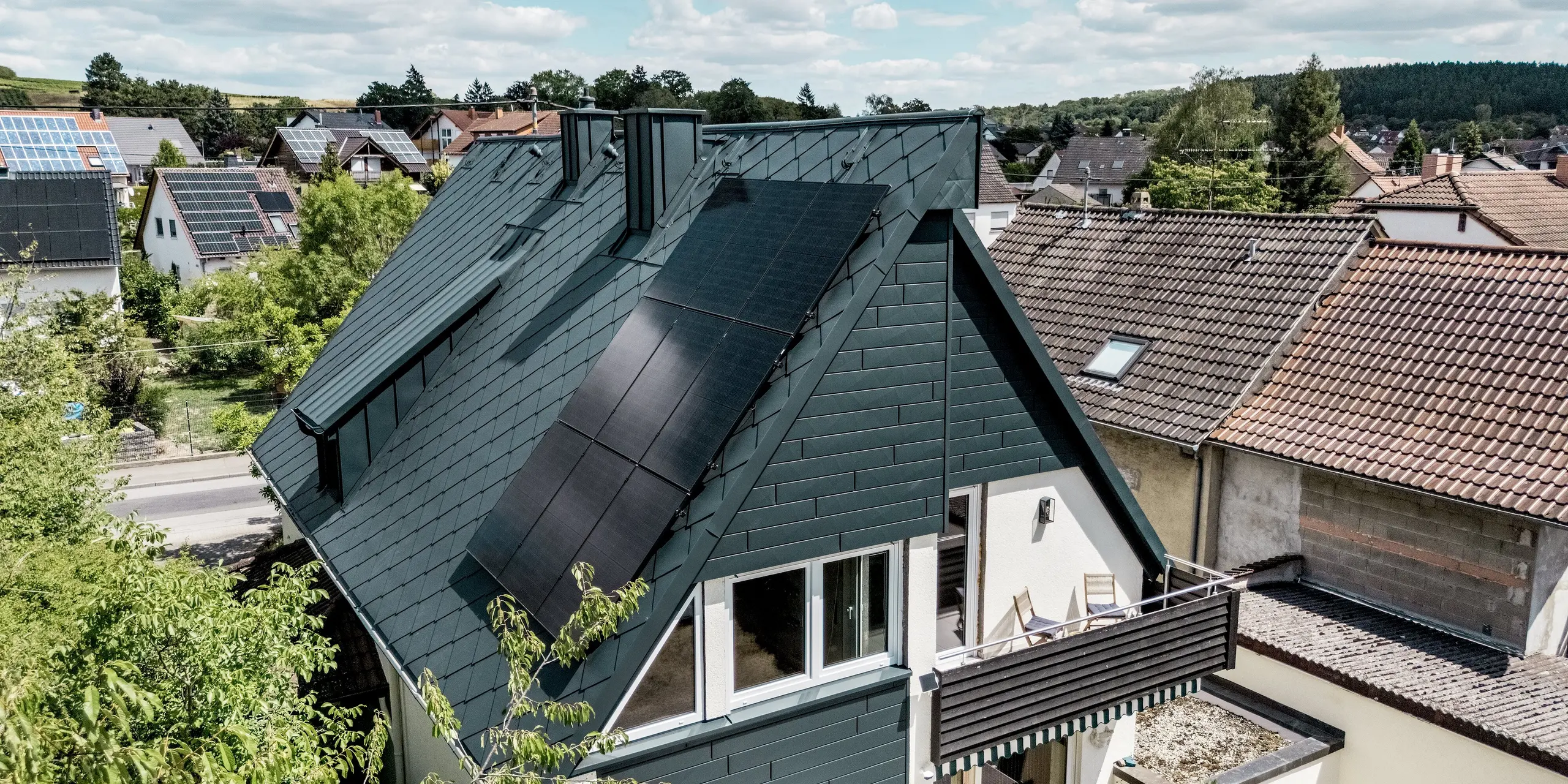 Luftaufnahme eines sanierten Einfamilienhauses in Hargesheim mit PREFA Dachraute 44 × 44 in P.10 Anthrazit. Die markante Dachstruktur harmoniert mit der Siding.X Giebelfassade und den mit PREFALZ verkleideten Gauben. Großflächige Photovoltaikmodule, befestigt mit dem PREFA Solarmontagesystem, ergänzen die langlebige, leichte und witterungsbeständige Aluminium-Dachlösung zu einem modernen Gesamtbild.