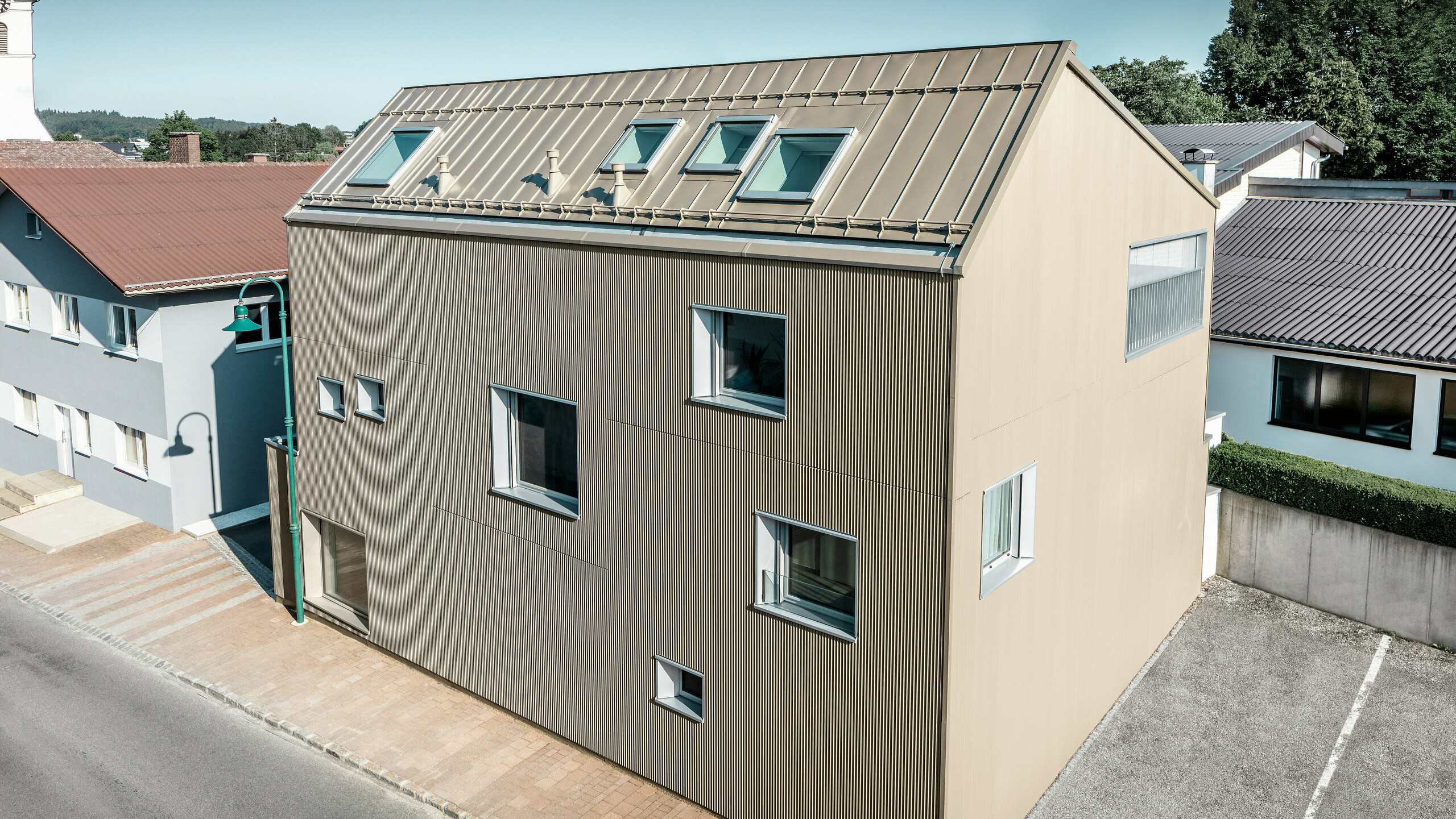 Vogelperspektive auf ein Einfamilienhaus in Frankenburg, Österreich, das hervorsticht durch seine PREFA Fassade in Form des Zackenprofils und einem PREFALZ-Dach, beide in abgestimmtem Bronze-Farbton. Die Fassade zeigt eine markante Textur, die das natürliche Licht einfängt und für ein lebendiges Schattenspiel sorgt. Mehrere Dachfenster lassen natürliches Licht ins Innere und unterstreichen die funktionale und ästhetische Integration moderner Architekturelemente. Das Haus fügt sich durch sein zeitgemäßes Design harmonisch in die typisch österreichische Dorf-Umgebung ein und setzt gleichzeitig einen kontrastreichen Akzent gegenüber den umliegenden traditionellen Gebäuden.