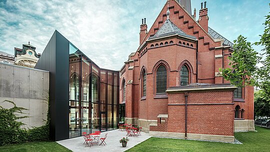 Das Bild zeigt den beeindruckenden Neubau, der die historische Rote Kirche mit der modernen Forschungsbibliothek in Olmütz verbindet. Die Perspektive zeigt die Rückseite des Gebäudes mit der einladenden Terrasse. Die elegante Fassade aus schwarzen PREFABOND Aluminium Verbundplatten setzt einen markanten, modernen Akzent und kontrastiert stilvoll mit dem roten Backstein der Kirche. Die hohe Präzision der Montage und die Verwendung langlebiger Materialien wie Aluminium gewährleisten eine harmonische Verbindung von Tradition und Innovation. Diese fortschrittlichen Platten sind nicht nur ästhetisch ansprechend, sondern auch äußerst funktional und langlebig, wodurch das Projekt zu einem Meisterwerk der modernen Architektur wird.