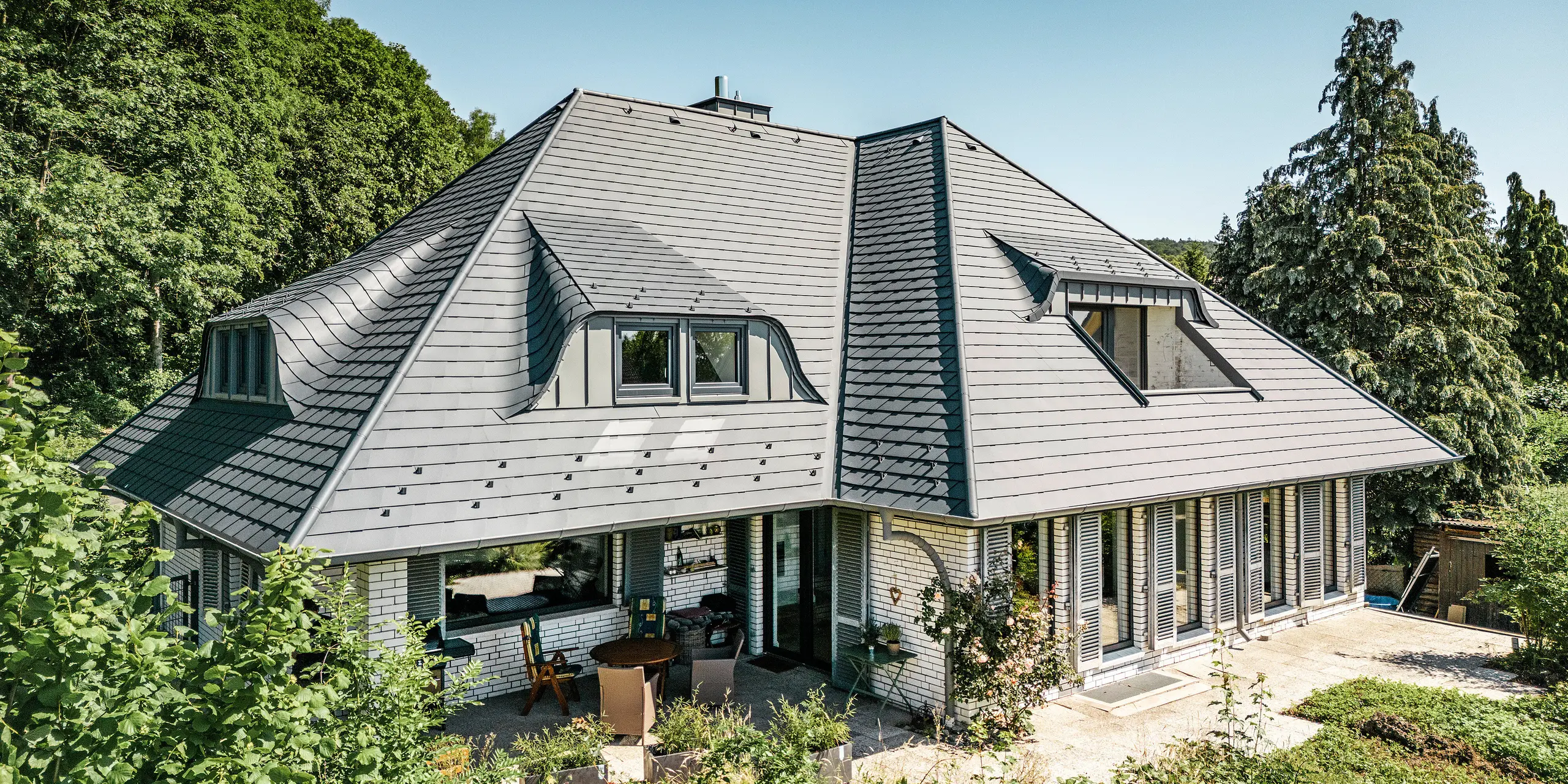 Moderne eengezinswoning in Einbeck, Duitsland, met PREFA dakspanen in P.10 antraciet. De duurzame PREFA dakspanen van aluminium staan bekend om hun kwaliteit en lange levensduur. Het dakontwerp van het gebouw benadrukt de individuele architectuur met puntgevels, die worden geaccentueerd door de flexibiliteit van de PREFA dakspanen. De dakafwatering in de vorm van de PREFA afvoerpijp en de hanggoot is eveneens in antraciet uitgevoerd. Rondom de eengezinswoning ligt een royale en goed onderhouden tuin met een terras. Achter het huis ligt een bos dat samen met de blauwe lucht een natuurlijke sfeer creëert.
