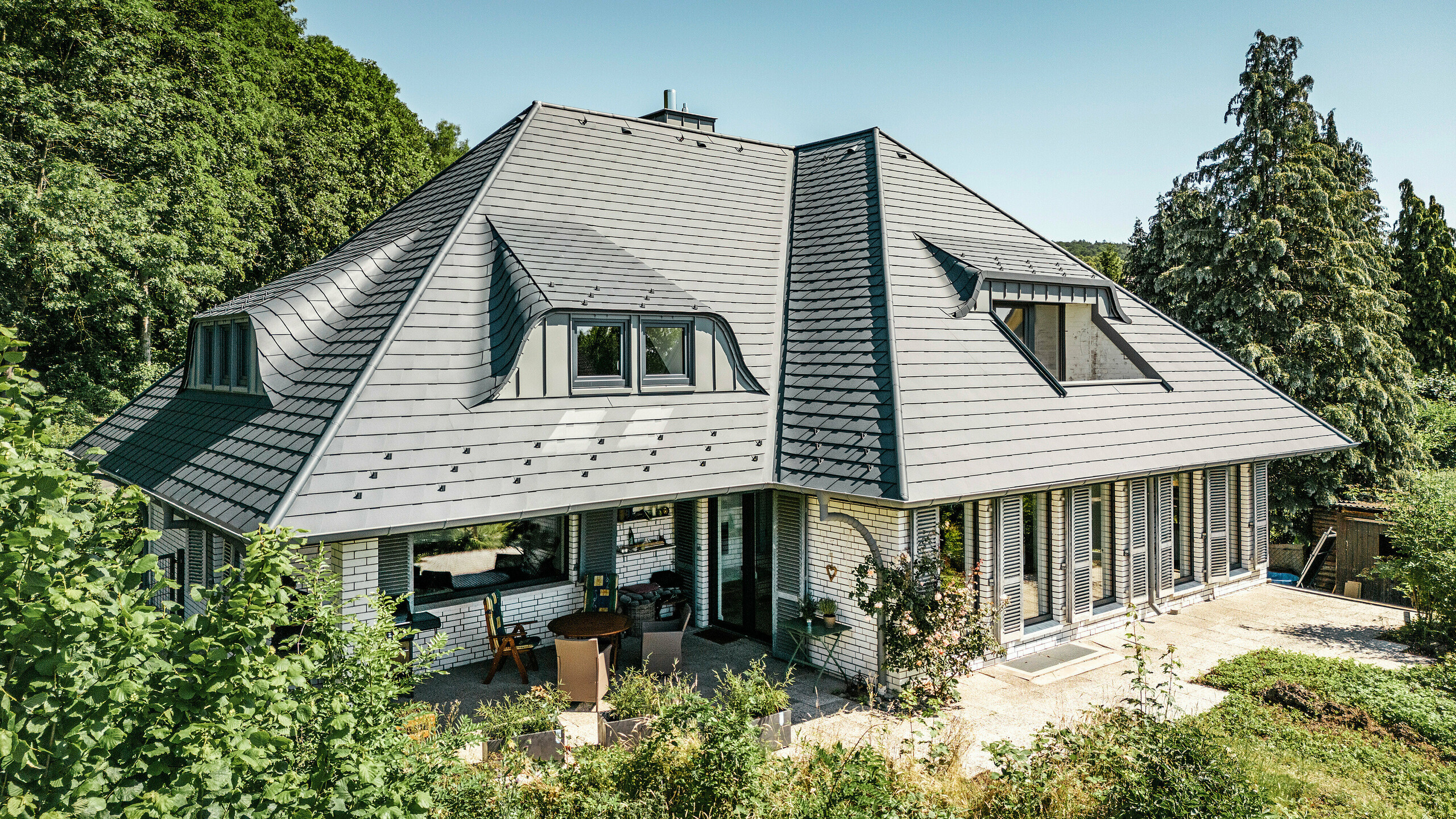 Moderne eengezinswoning in Einbeck, Duitsland, met PREFA dakspanen in P.10 antraciet. De duurzame PREFA dakspanen van aluminium staan bekend om hun kwaliteit en lange levensduur. Het dakontwerp van het gebouw benadrukt de individuele architectuur met puntgevels, die worden geaccentueerd door de flexibiliteit van de PREFA dakspanen. De dakafwatering in de vorm van de PREFA afvoerpijp en de hanggoot is eveneens in antraciet uitgevoerd. Rondom de eengezinswoning ligt een royale en goed onderhouden tuin met een terras. Achter het huis ligt een bos dat samen met de blauwe lucht een natuurlijke sfeer creëert.