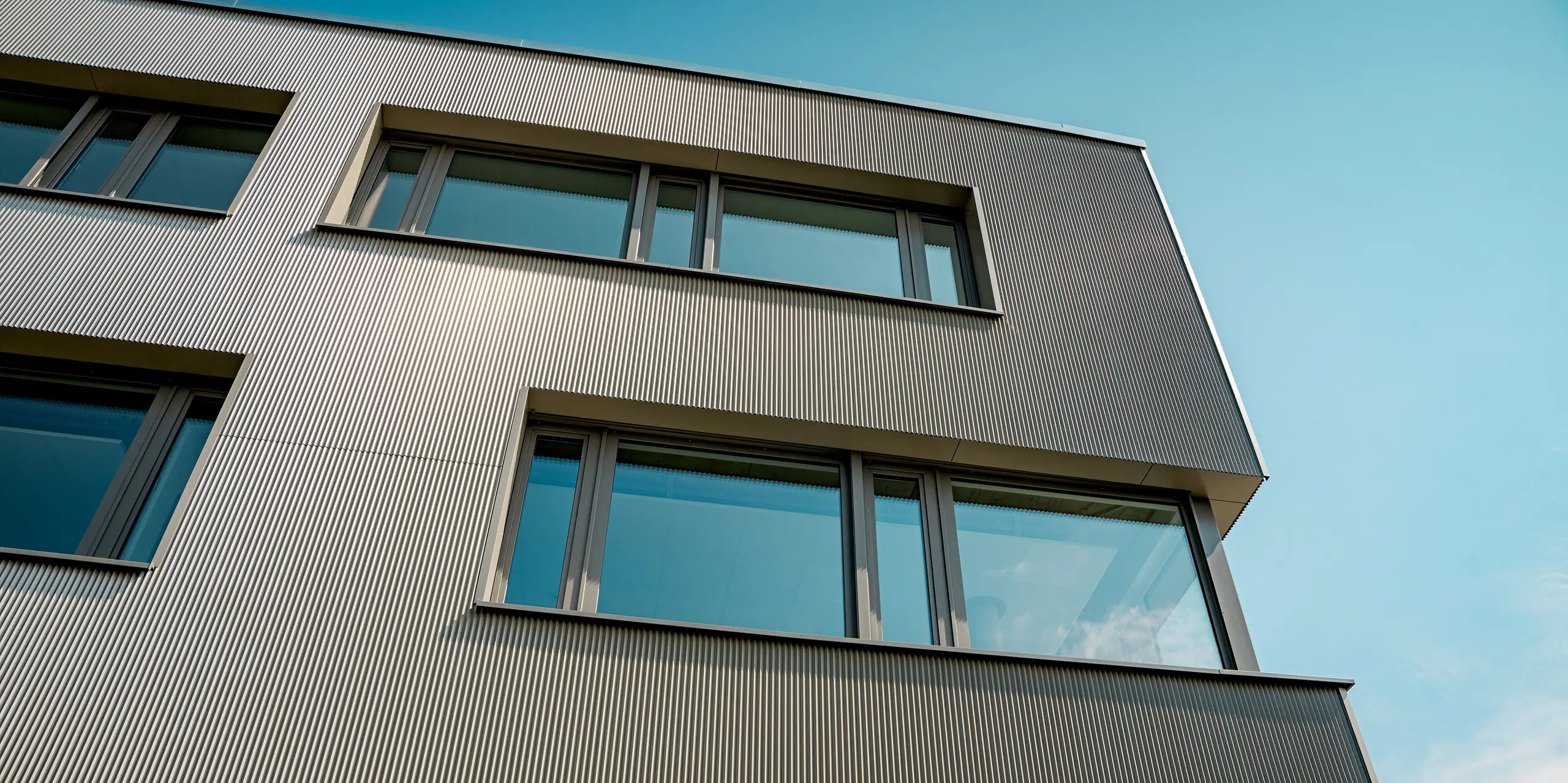 L'image présente un gros plan de la façade en aluminium du bâtiment Festo à Langenhagen, revêtue de profils extrudés triangle PREFA. La structure verticale des profils confère à la façade un aspect texturé saisissant, rendant le bâtiment à la fois moderne et dynamique. La pureté des lignes et l'intégration précise des fenêtres sont particulièrement remarquables, créant un équilibre harmonieux entre architecture et fonctionnalité. L'utilisation des produits PREFA, robustes et durables, garantit une façade esthétique et résistante aux intempéries, répondant aux exigences les plus élevées en matière de design et de développement durable.