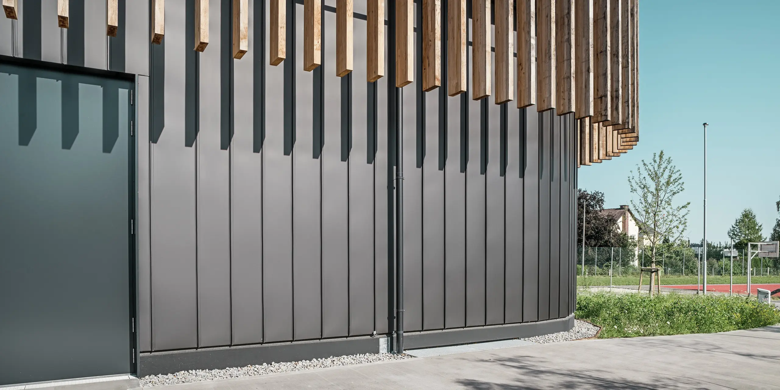 Gros plan du rez-de-chaussée du Team 7 World à Ried im Innkreis : la façade PREFALZ noir-brun forme une image globale harmonieuse avec les éléments verticaux en bois. Le revêtement robuste en aluminium protège des intempéries et impressionne par sa qualité durable et sans entretien. Le jeu d'ombre et de lumière souligne les lignes claires et l'architecture moderne du complexe immobilier.