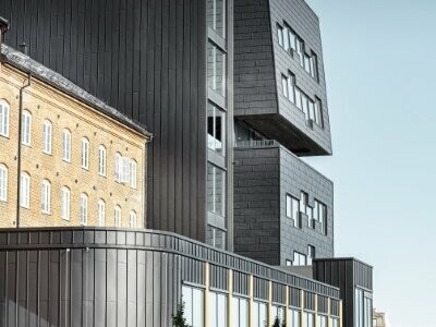 Seitenansicht des Gebäudes in Norwegen. Vor dem Gebäude sind Bäume zu sehen. Das Foto wurde bei strahlendem Sonnenschein aufgenommen. Das Gebäude wurde mit den FX.12 Fassadenpaneelen und PREFALZ in der Farbe P.10 verkleidet und steht leicht im Schatten.