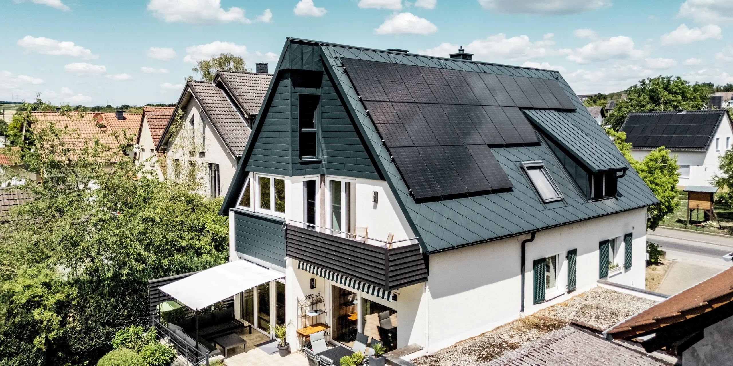 Luftaufnahme eines modern sanierten Einfamilienhauses in Hargesheim mit PREFA Dachraute 44 × 44 in P.10 Anthrazit. Großflächige Photovoltaikmodule und eine mit PREFALZ verkleidete Dachgaube setzen markante Akzente. Die Siding.X Giebelfassade ergänzt das harmonische Gesamtbild. Die leichten, langlebigen und witterungsbeständigen Aluminiumprodukte verbinden moderne Architektur mit nachhaltiger Energiegewinnung.