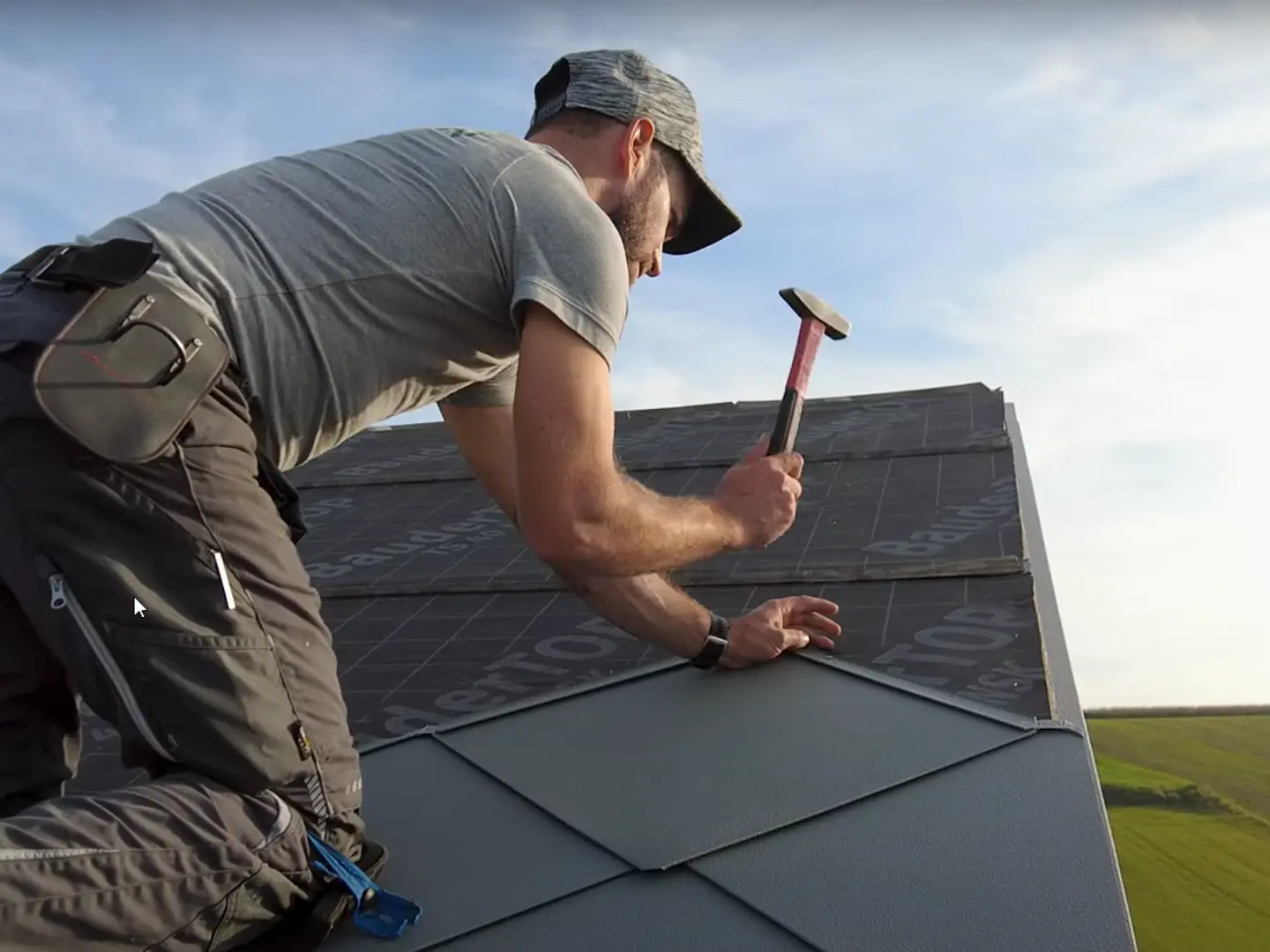 Loodgieter in actie: met een hamer legt de eigenaar van het bedrijf ‘Der Blechmann’ nauwkeurig de PREFA daklosange 44 × 44. De precisie bij de installatie is te zien aan de reeds gelegde daklosangen aan de onderkant van de foto. De man draagt een pet en knielt met zijn rug naar de camera op het dak. Op de achtergrond zijn de heldere lucht en een groen landschap te zien. De harmonieuze achtergrond onderstreept de symboliek van de foto: duurzaam en langdurig vakmanschap met hoogwaardige PREFA aluminiumproducten.