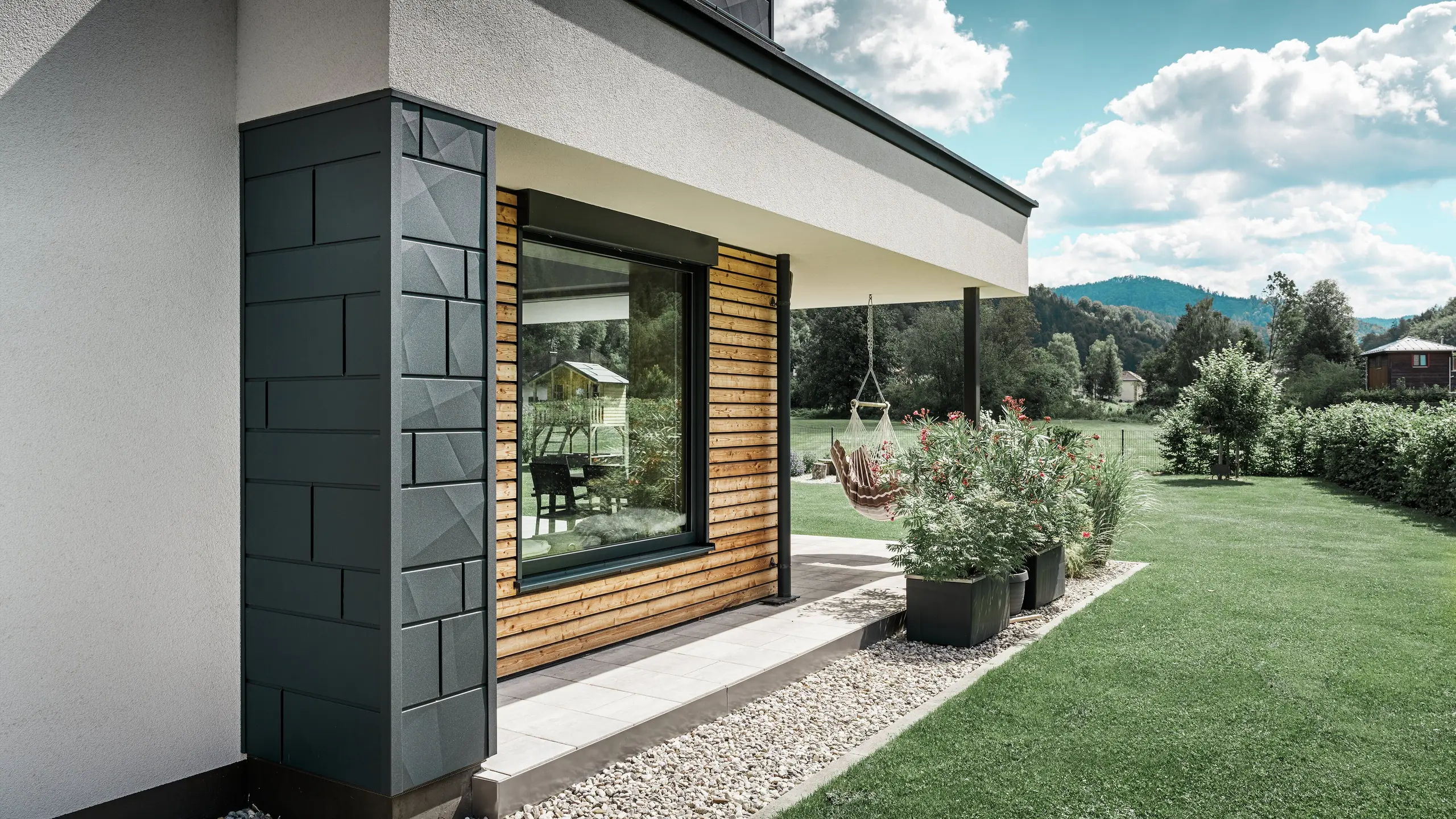 Gedeeltelijk aanzicht van een nieuw gebouwd vrijstaand huis. Hier werden verschillende materialen voor de gevel gebruikt. De wit gepleisterde gevel wordt geaccentueerd door PREFA aluminium Siding.X in antraciet en horizontale houten panelen, die een groot raam omlijsten. Op het terras hangt een hangstoel.