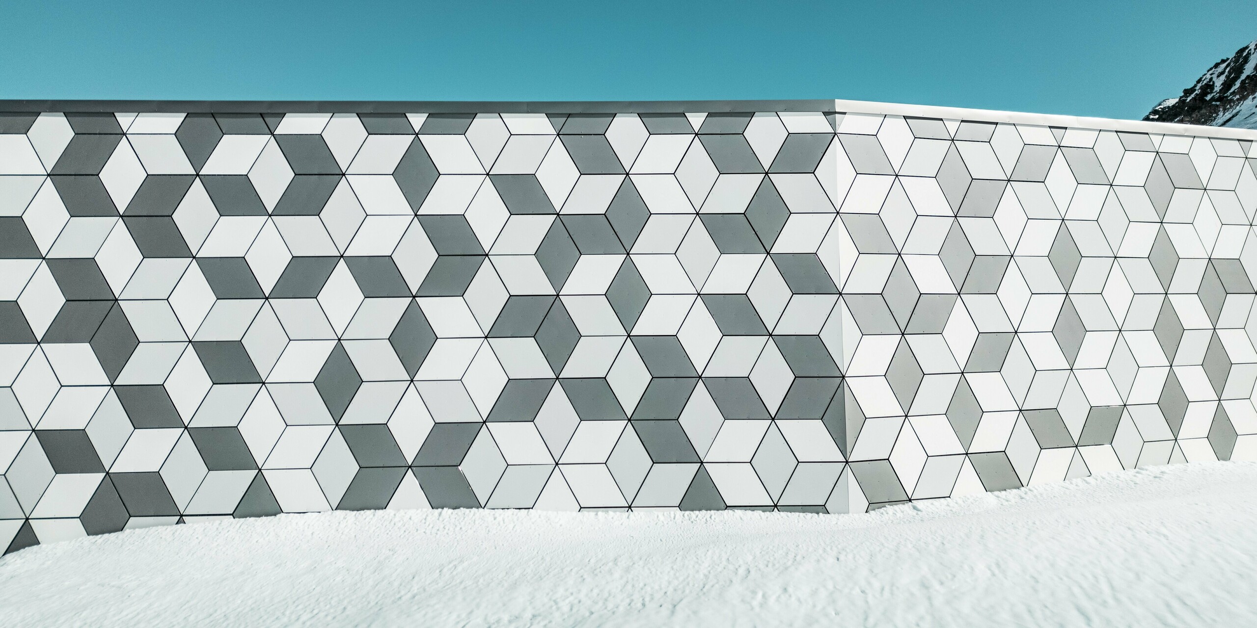 Ce gros plan de la façade de la station de téléphérique Jandri aux Deux Alpes illustre la disposition précise des losanges réalisés en panneaux composites en aluminium PREFABOND. Ces losanges forment un motif saisissant. Grâce à une palette de couleurs harmonieuse, déclinée en nuances de blanc et de gris, la façade s'intègre parfaitement au paysage alpin enneigé. Robustes et résistants aux intempéries, ces panneaux constituent une solution durable et esthétique pour les conditions climatiques extrêmes en montagne.
