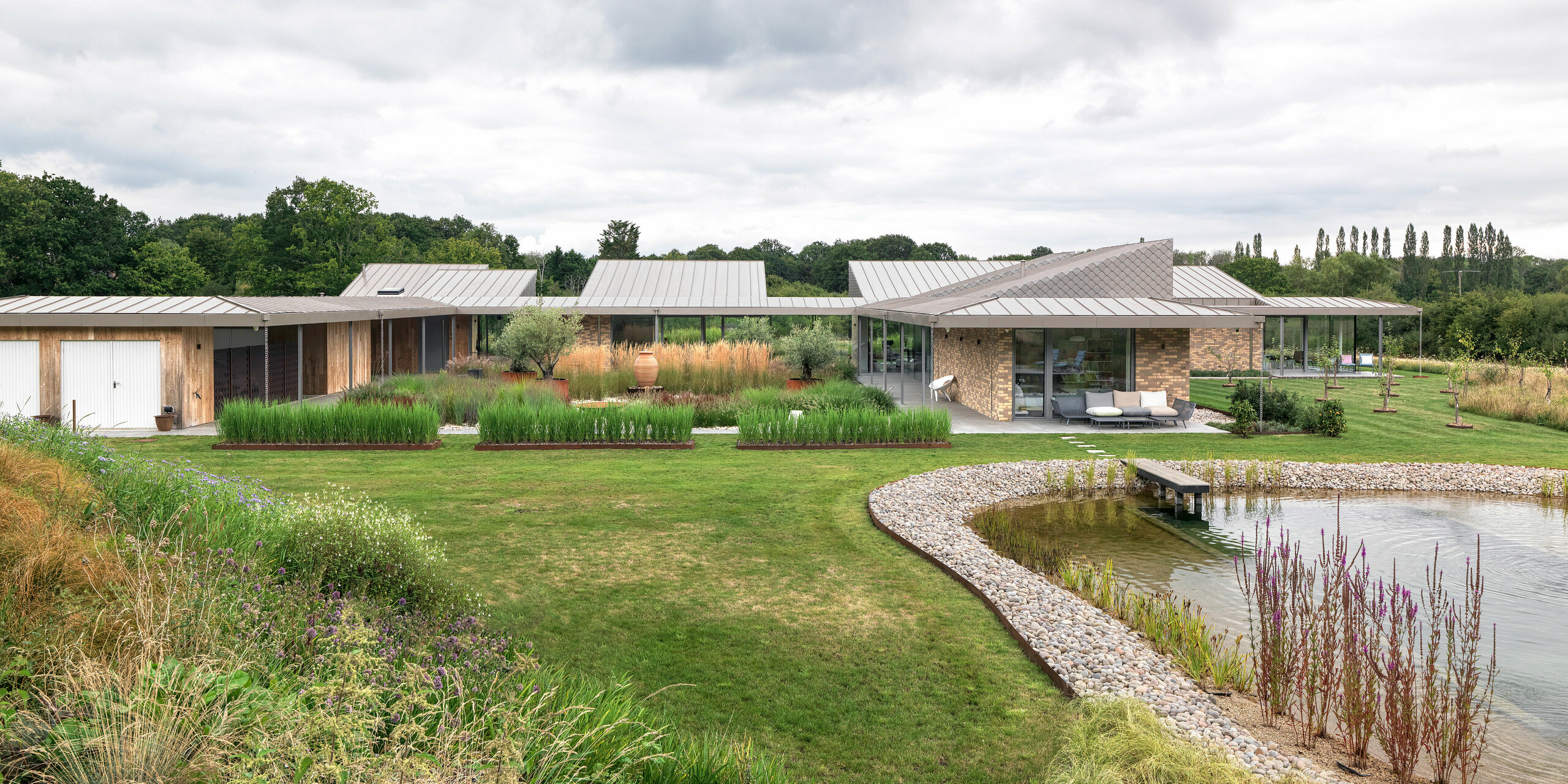 Vue panoramique de cet immeuble résidentiel à l'architecture sophistiquée situé dans le West Sussex, avec sa grande toiture en aluminium PREFALZ couleur P.10 bronze et ses losanges de façade PREFA harmonieusement intégrés à certains endroits de la façade. Ce bâtiment moderne s'étend sur toute la propriété, plat et spacieux, au cœur d'une végétation naturelle et avec un bassin naturel paysager au premier plan. Les robustes produits PREFA s'inscrivent dans le concept de construction durable et garantissent un aspect uniforme et durable.
