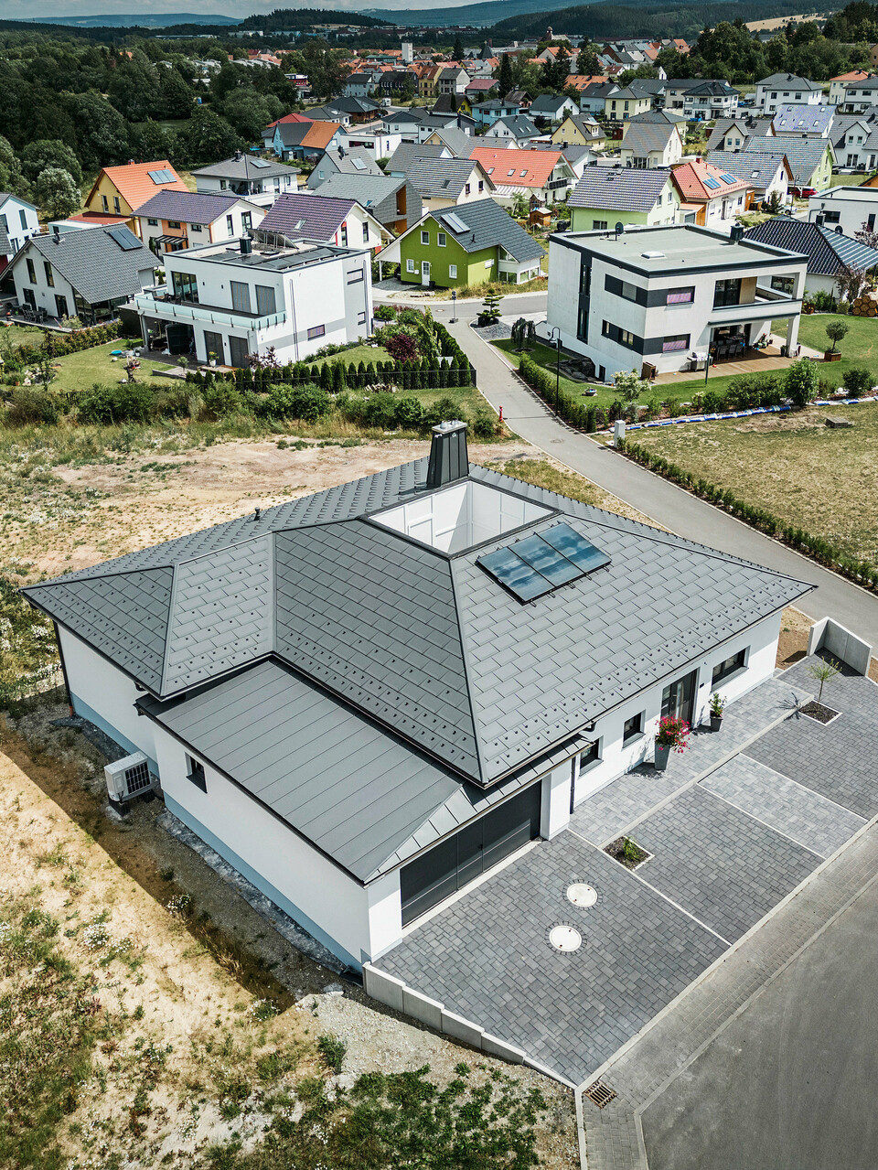 Moderne Architektur trifft Funktionalität: Ein Bungalow in Ilmenau, Thüringen, mit einem beeindruckenden PREFA Aluminiumdach bestehend aus der Dachplatte R.16 in P.10 Dunkelgrau. Die Dachlandschaft zeigt ein zentrales Atrium und einen markanten Schornstein mit dem charakteristischen PREFA Stier, ergänzt durch ein langlebiges PREFA Dachentwässerungssystem mit Ablaufrohr und Hängerinne. Die Garage fügt sich nahtlos in das Gesamtbild ein, verkleidet mit PREFALZ in passendem Dunkelgrau.