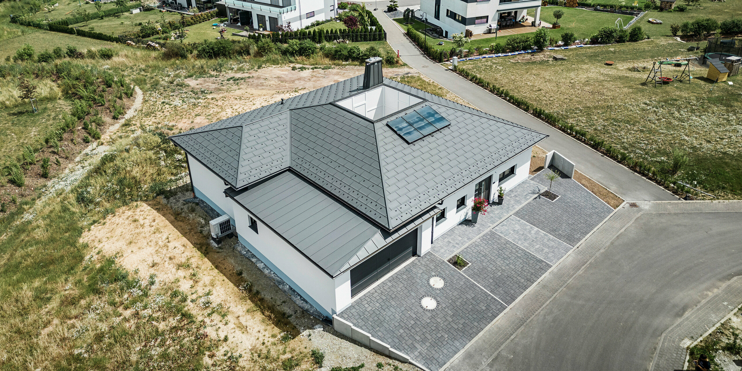 Moderne Architektur trifft Funktionalität: Ein Bungalow in Ilmenau, Thüringen, mit einem beeindruckenden PREFA Aluminiumdach bestehend aus der Dachplatte R.16 in P.10 Dunkelgrau. Die Dachlandschaft zeigt ein zentrales Atrium und einen markanten Schornstein mit dem charakteristischen PREFA Stier, ergänzt durch ein langlebiges PREFA Dachentwässerungssystem mit Ablaufrohr und Hängerinne. Die Garage fügt sich nahtlos in das Gesamtbild ein, verkleidet mit PREFALZ in passendem Dunkelgrau.