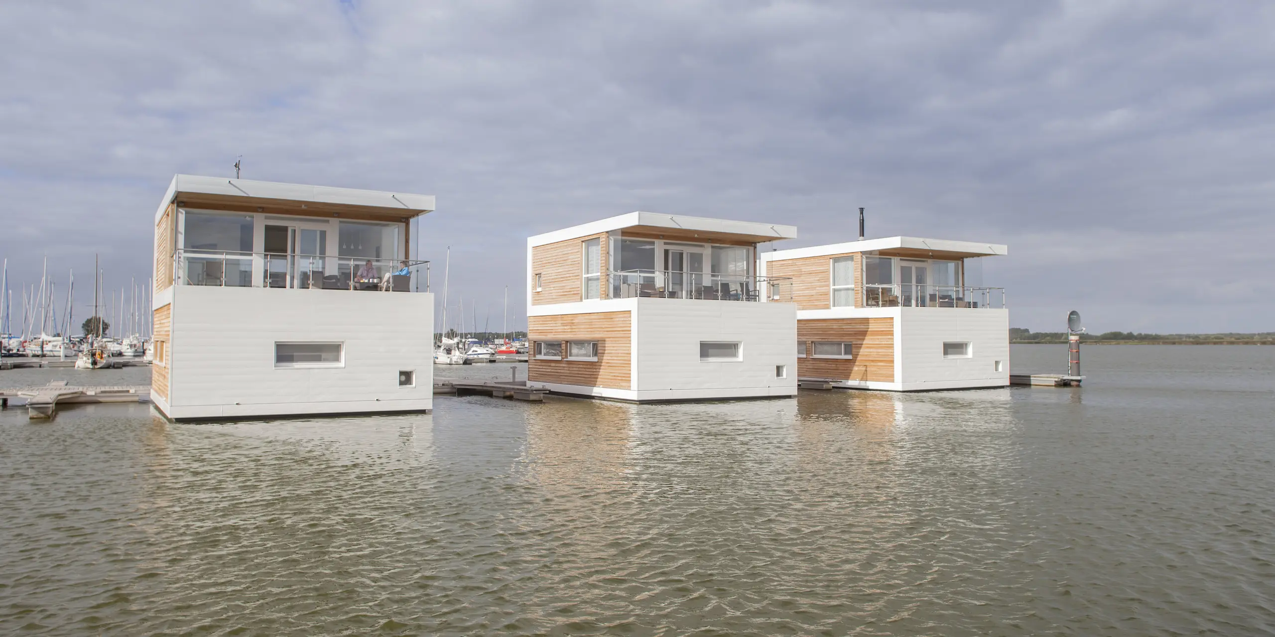 Drei moderne schwimmende Häuser in Stralsund liegen direkt im Wasser des Yachthafens und beeindrucken mit klarer, geradliniger Architektur. Die PREFA Sidings in P.10 Prefaweiß setzen markante Akzente und bilden einen eleganten Kontrast zu den warmen Holzfassaden. Die langlebigen, korrosionsbeständigen Aluminiumfassaden sind ideal für das maritime Umfeld und überzeugen durch ihre Witterungsbeständigkeit sowie Pflegeleichtigkeit.